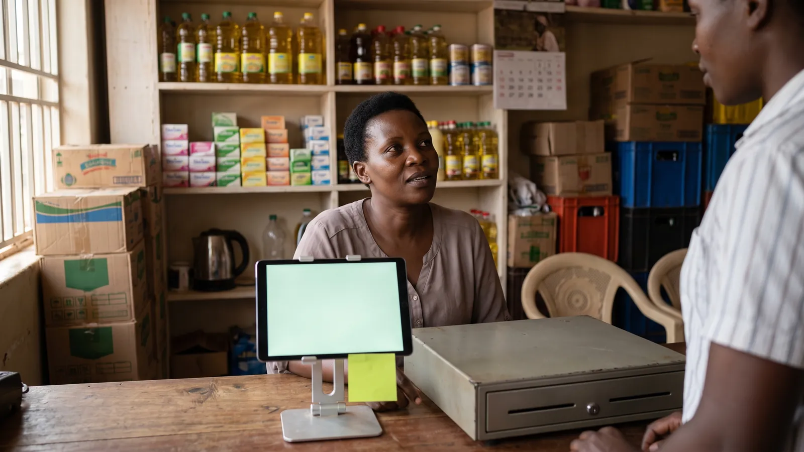 A shop owner at the counter of a small supermarket in Kampala, a tablet on a stand beside the cash drawer